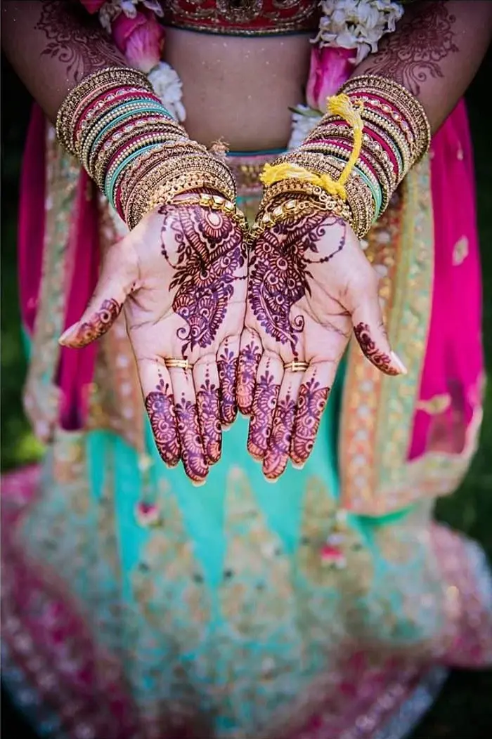 Bride displaying palm mehndi with gold bangles and floral garland, wearing teal and pink bridal lehenga
